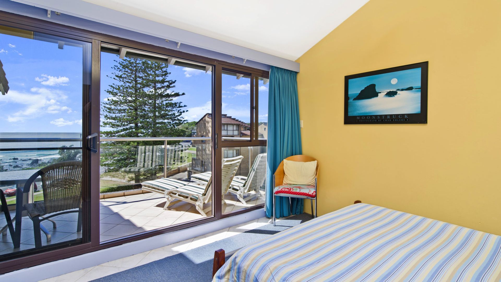 A room with a bed, a chair, a table, and a large sliding glass door leading to a balcony with a view of the ocean and a tree.
