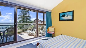 A room with a bed, a chair, a table, and a large sliding glass door leading to a balcony with a view of the ocean and a tree. - Beachpark 56, 58 Pacific Drive (Port Macquarie)