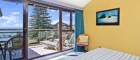 A room with a bed, a chair, a table, and a large sliding glass door leading to a balcony with a view of the ocean and a tree.