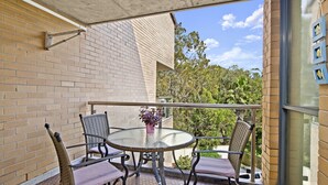 A balcony with a round glass table and four wicker chairs, a potted plant, and a view of trees and sky. - Beachpark 56, 58 Pacific Drive (Port Macquarie)