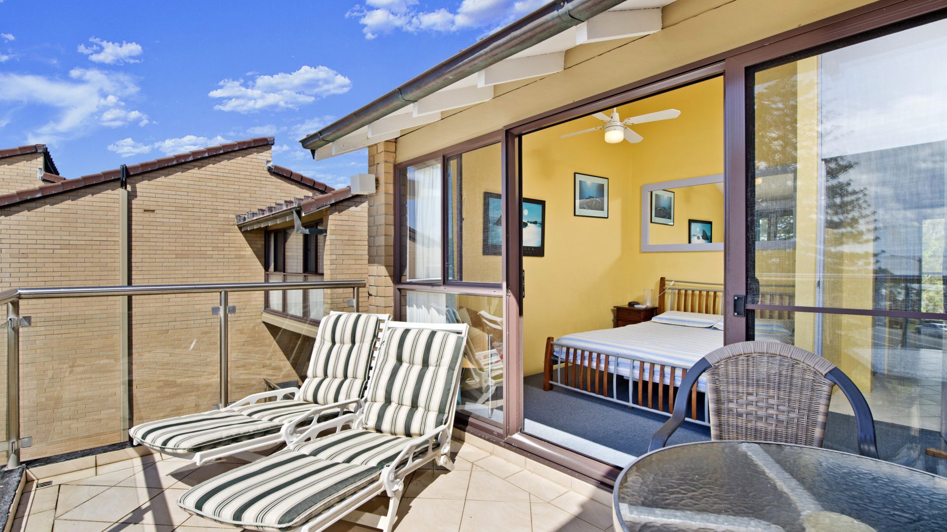 A balcony with two lounge chairs and a table, a bedroom with a bed and a nightstand, and a large window with a view of the sky.