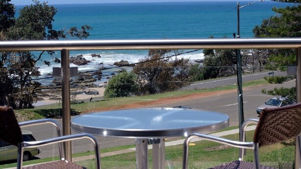 A balcony with a metal railing, a round table, and two chairs. The view includes a coastal road, a beach with rocks, and the ocean. - Beachpark 56, 58 Pacific Drive (Port Macquarie)