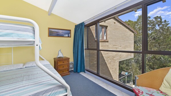 A room with bunk beds, a small dresser, a fan, and a large window with a view of a brick building and trees.