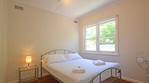 A single bed with white linens, a bedside table with a lamp, a window with a view of greenery, and a standing fan.