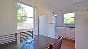 A compact kitchen with white appliances, a dining table with chairs, and windows with a view of greenery.