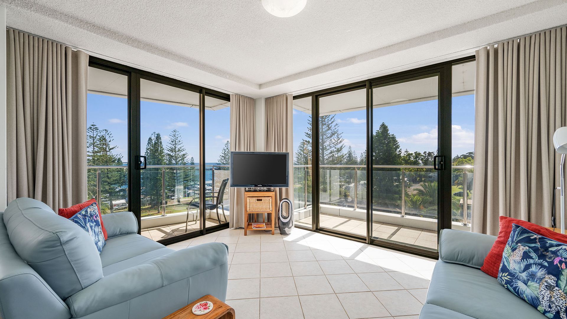 A spacious living room with a large flat-screen TV, two sofas, and a balcony with a view of trees and a body of water.