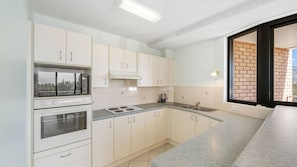 A modern kitchen with white cabinetry, a built-in oven, a microwave, a kettle, and a window with a view of a brick wall.
