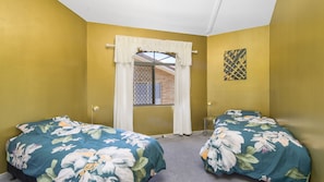 A room with two beds, a window with a white curtain, a small table with a lamp, and a wall-mounted artwork.
