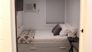 A compact bedroom with a single bed, a small window with blinds, a wall-mounted air conditioning unit, and a wooden floor. - Mini farmhouse style container Homes #4 (Orderville)