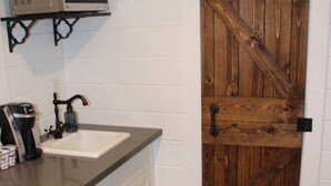 A kitchen with a white countertop, a sink, a microwave, and a wooden barn-style door. - Mini farmhouse style container Homes #4 (Orderville)