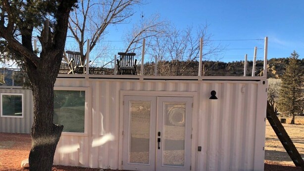 A small, white, single-story building with a door and windows, surrounded by gravel and trees. - Mini farmhouse style container Homes #4 (Orderville)