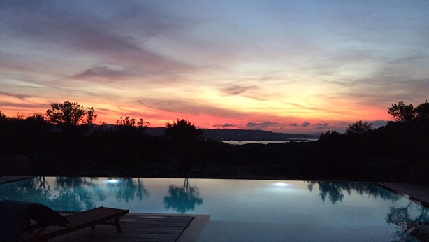 Une piscine à débordement avec une terrasse en bois et des chaises longues, entourée d’arbres et de collines, au coucher du soleil.