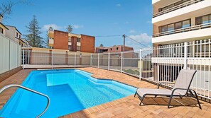 Una piscina con valla blanca, una silla de descanso y un edificio al fondo.