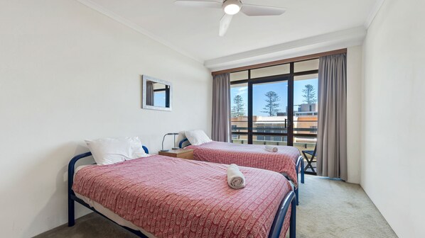 A hotel room with two beds, a ceiling fan, a window with curtains, and a view of buildings and trees.