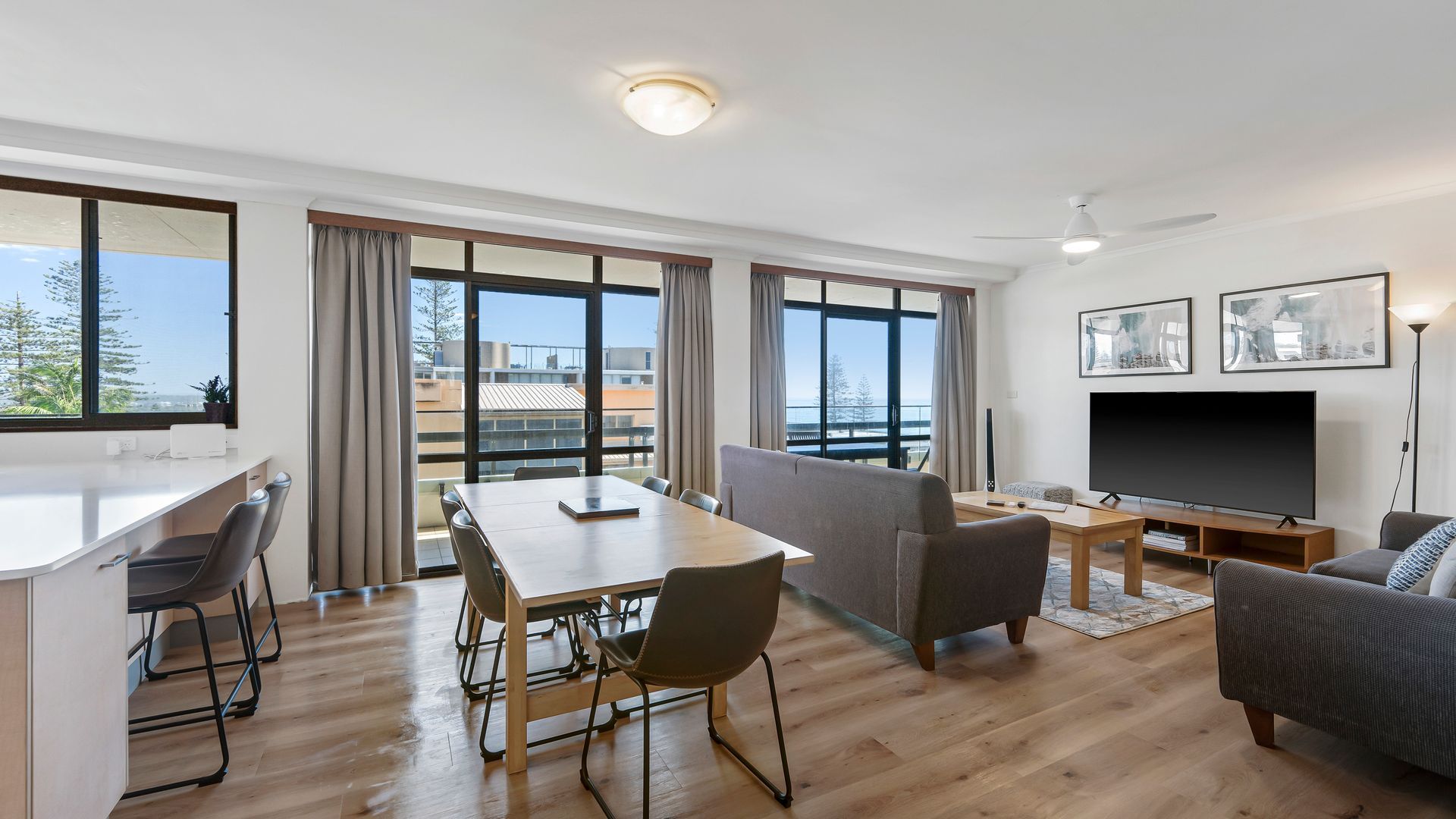 A modern living room with a flat-screen TV, a dining area with a table and chairs, and large windows offering a view of the outdoors.