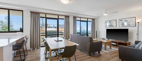 Ein modernes Wohnzimmer mit Fernseher, einer Essecke mit Tisch und Stühlen sowie großen Fenstern mit Blick ins Freie.