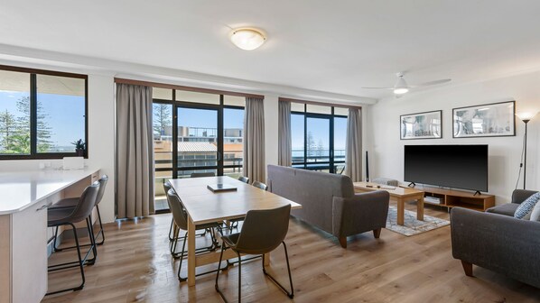 Una sala moderna con un televisor de pantalla plana, un comedor con mesa y sillas, y ventanas amplias que ofrecen vistas al exterior.