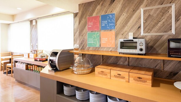 A breakfast bar with a microwave, toaster, and bread boxes. There are plates stored under the counter and a window with blinds in the background. - Comfort Inn Kashima (Kamisu)