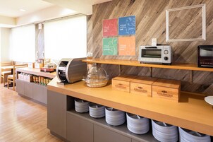 A breakfast bar with a microwave, toaster, and bread boxes. There are plates stored under the counter and a window with blinds in the background.