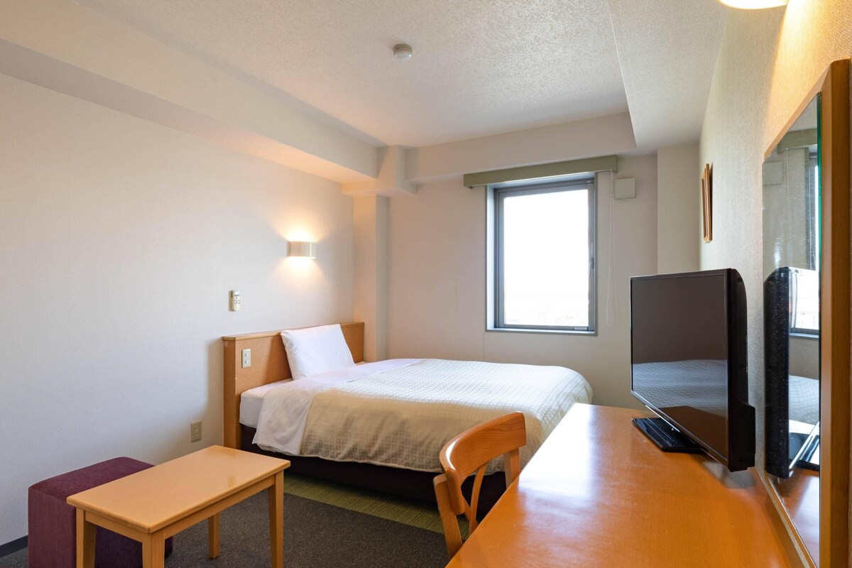 Ein Hotelzimmer mit einem Bett, einem Schreibtisch, einem Sessel, einem Fernseher und einem Fenster.