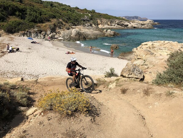 A mountain biker on a dirt path near a beach with rocky terrain and a clear view of the ocean.