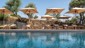 A poolside area with lounge chairs and umbrellas, a stone wall, and trees in the background.