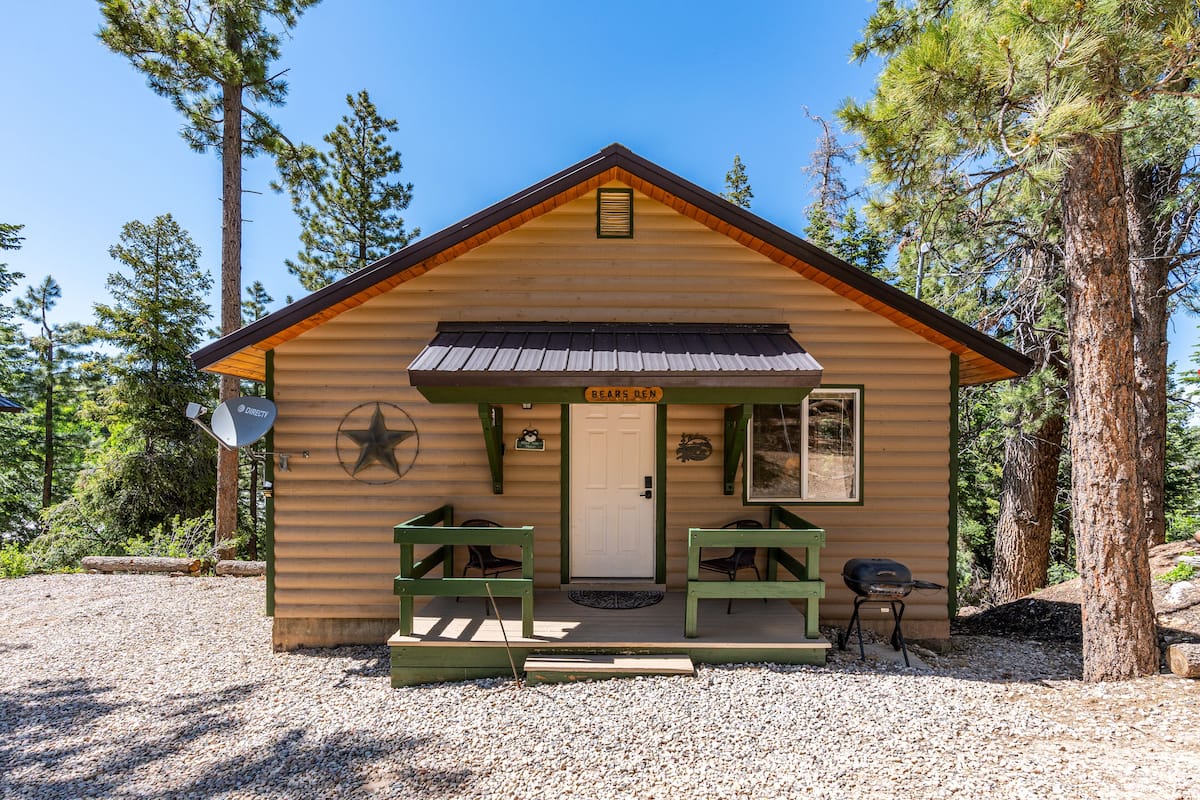 Une petite cabane avec une façade en bois, une porte d’entrée, deux bancs, un barbecue et une antenne parabolique.