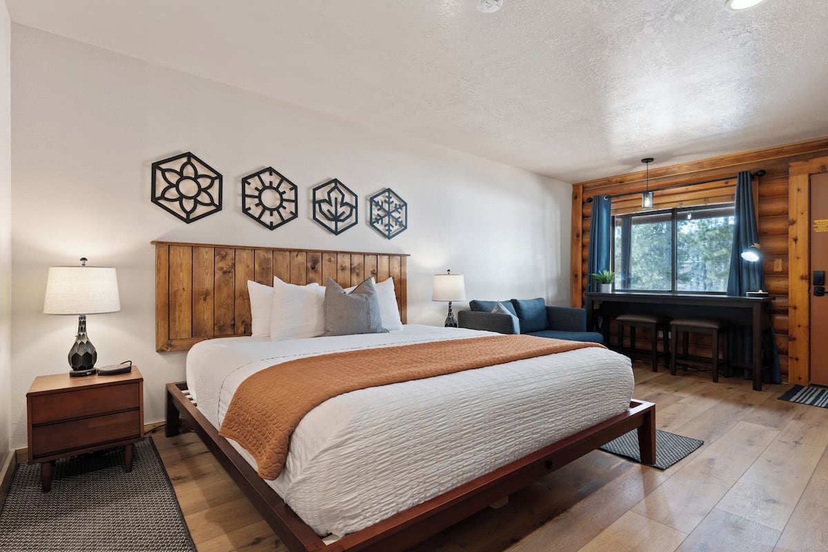 Une chambre à coucher avec une tête de lit en bois, un grand lit avec une literie blanche, un plaid marron et un bureau avec une chaise et des tabourets de bar.