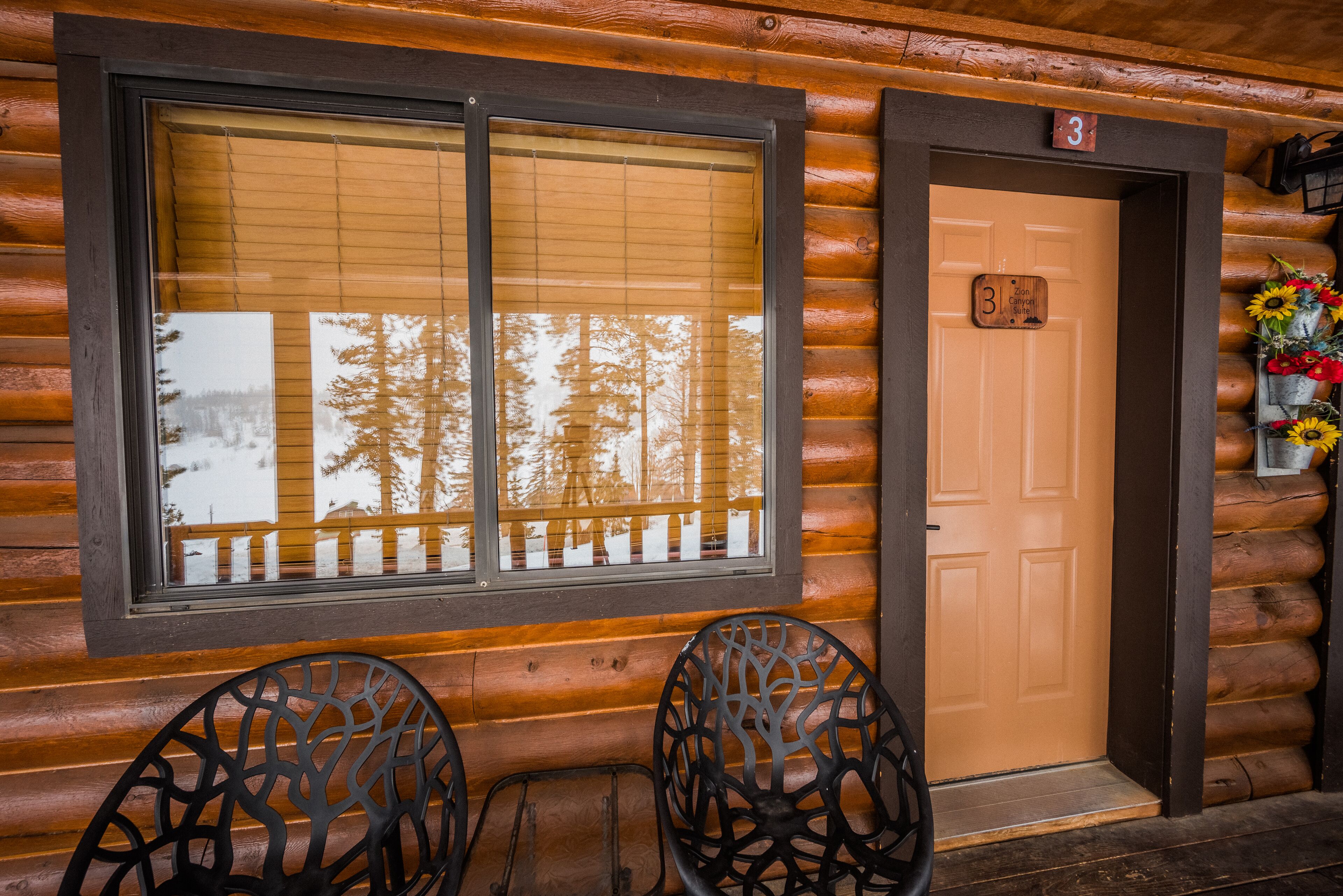 A log cabin with a large window, a door with a number, and two chairs outside.