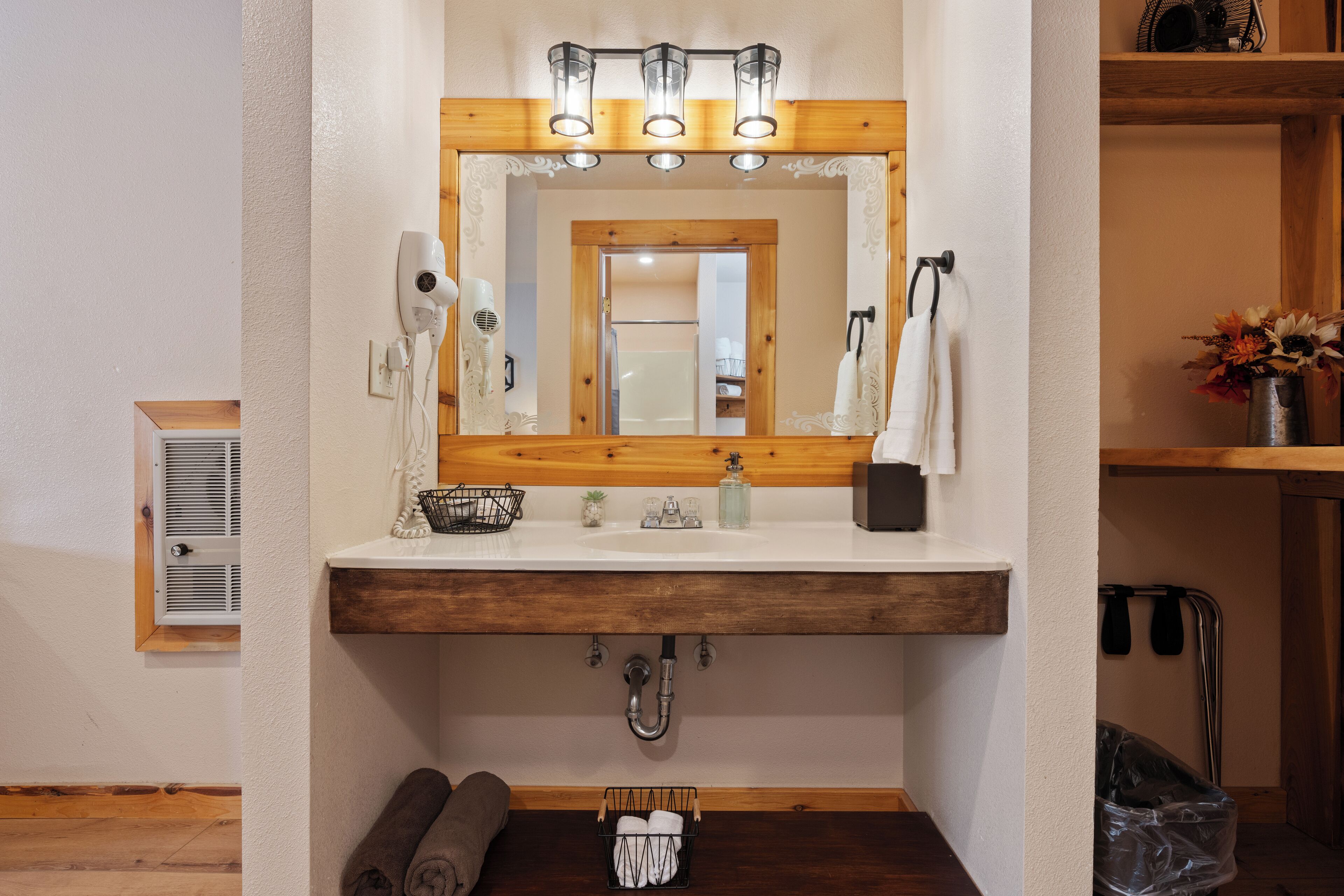 A bathroom with a wooden vanity, a mirror with lights, a towel rack, and a small plant.