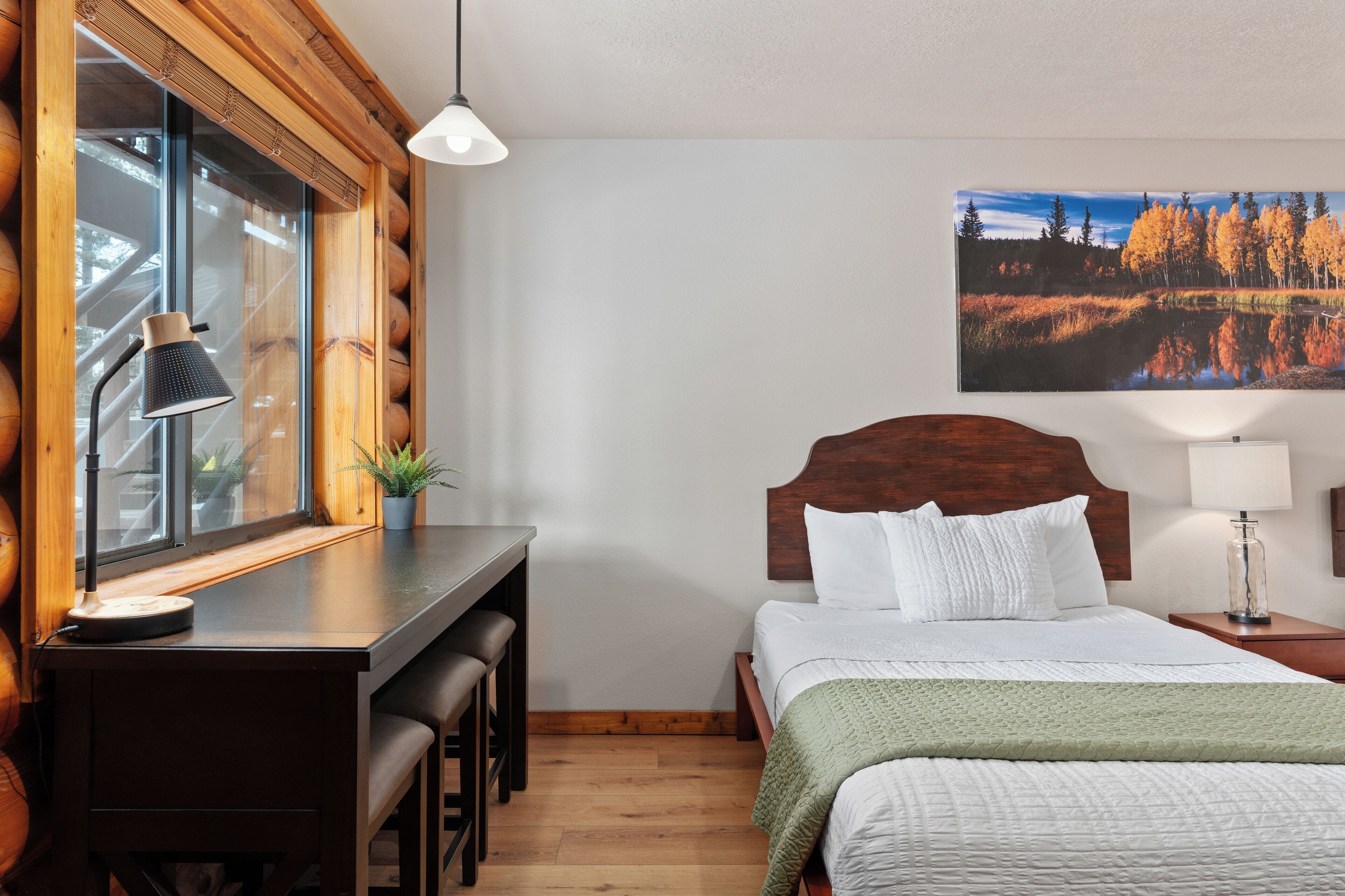 A bedroom with a wooden headboard, a desk with a lamp, a bed with white bedding, and a large window with a view of trees.