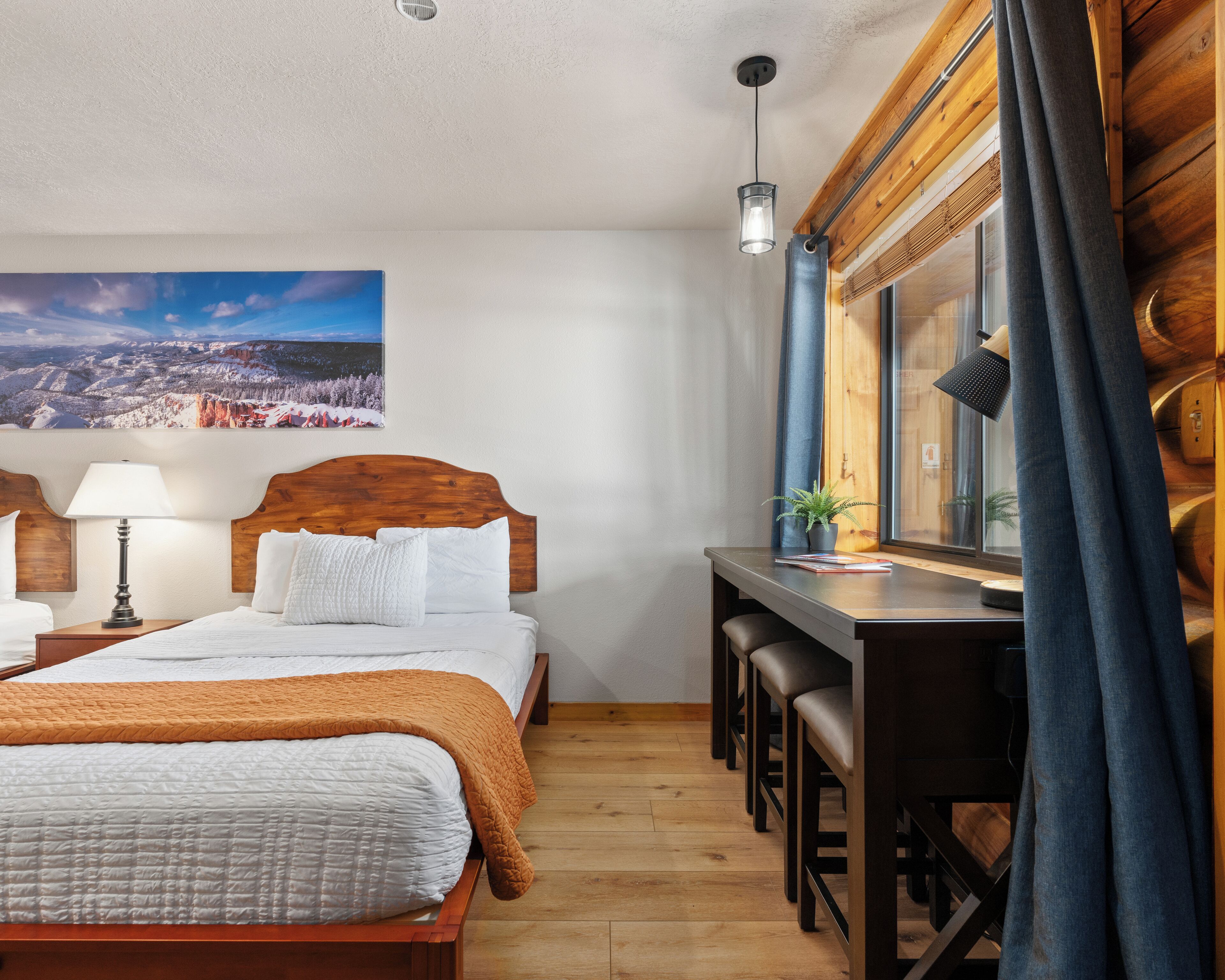 A hotel room with a wooden bed, a desk with chairs, a window with curtains, a lamp, and a painting on the wall.