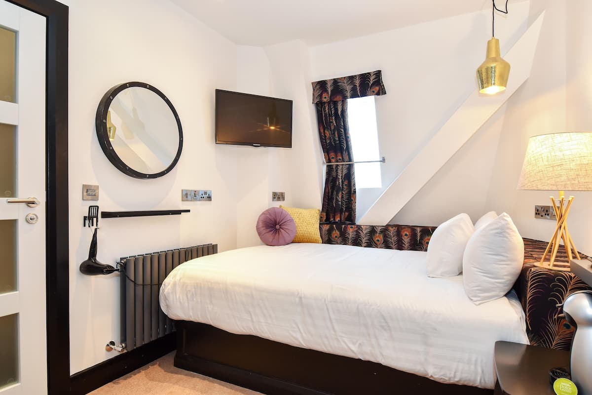 A bedroom with a bed, a television, a mirror, a window with curtains, a radiator, and a lamp.