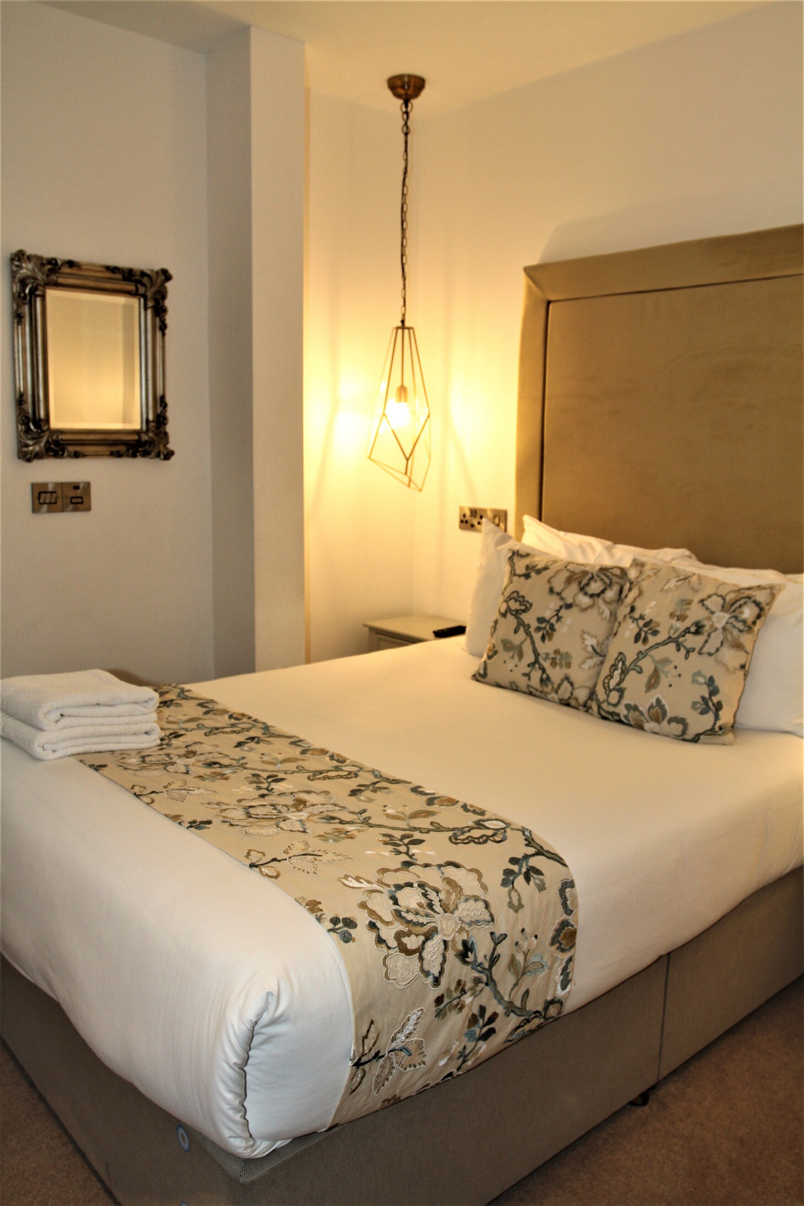 A neatly made bed with a floral bedspread, a bedside lamp, and a mirror on the wall.