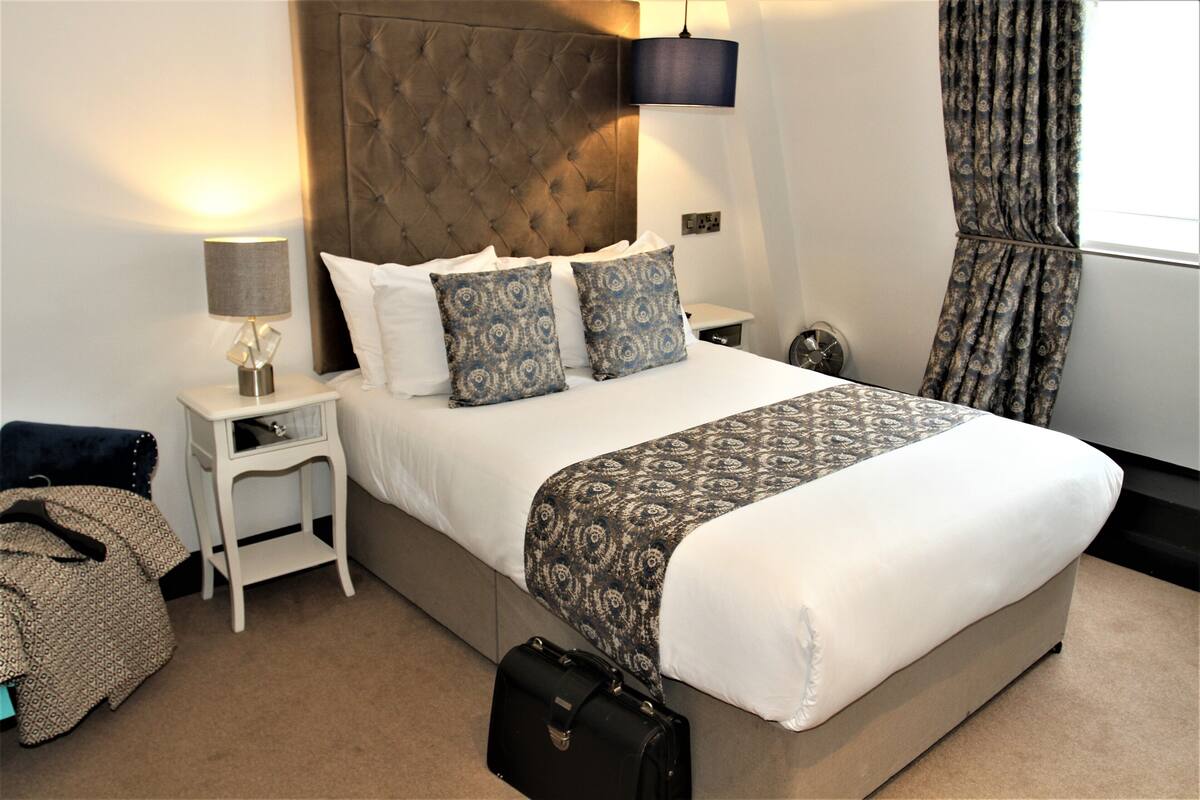 A neatly made bed with a patterned bedspread, a bedside lamp, and a black suitcase on the floor.