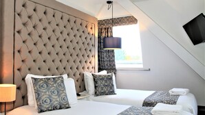 A hotel room with two beds, a large tufted headboard, a window with curtains, a flat-screen TV mounted on the wall, and a lamp on the side table. - The Lane - Boutique Residence (Galway)