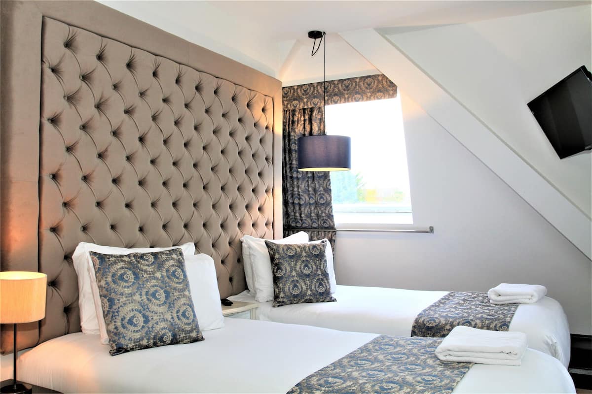 A hotel room with two beds, a large tufted headboard, a window with curtains, a flat-screen TV mounted on the wall, and a lamp on the side table.