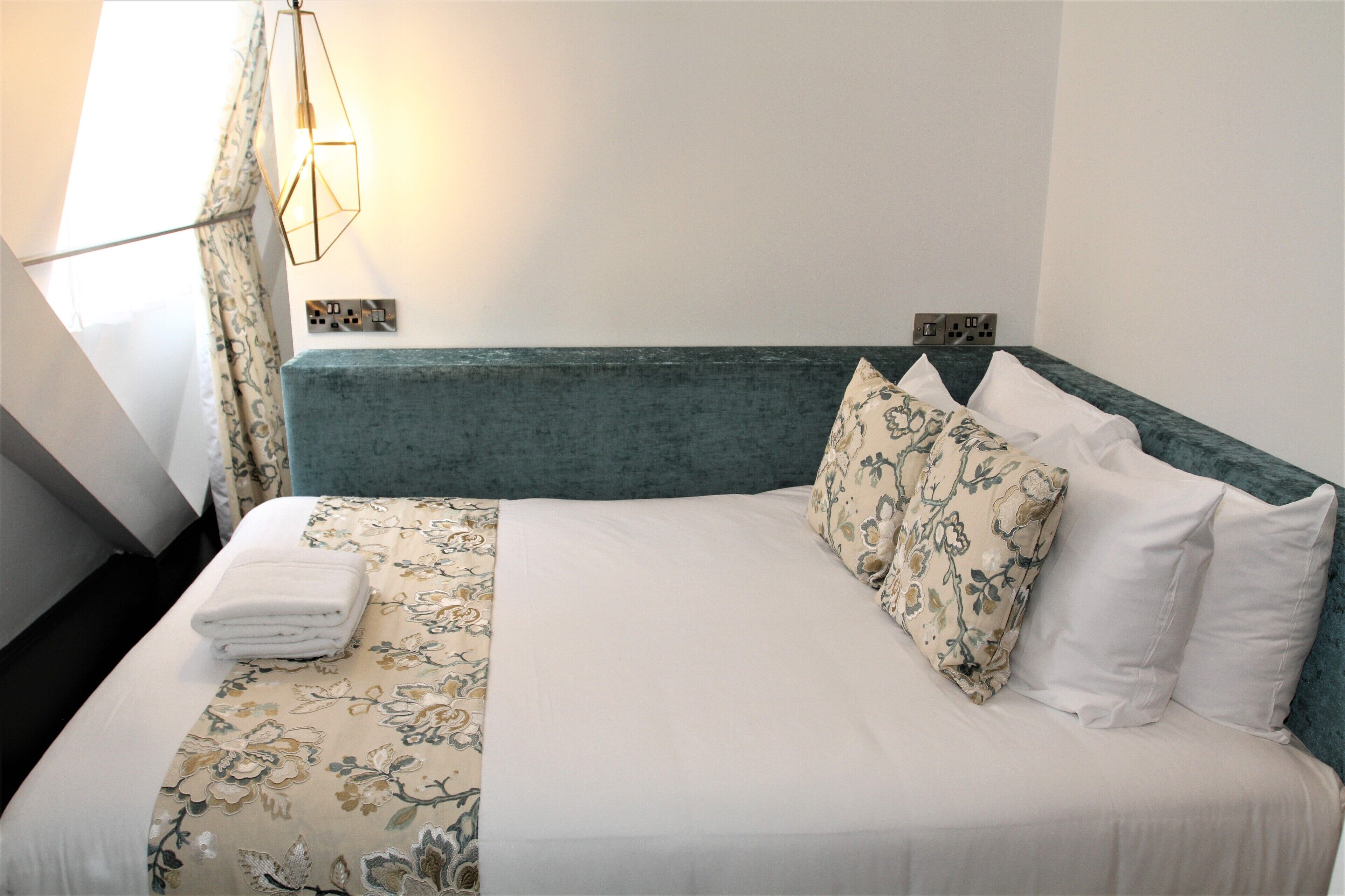 A neatly made bed with floral bedding, a teal headboard, and a bedside lamp.