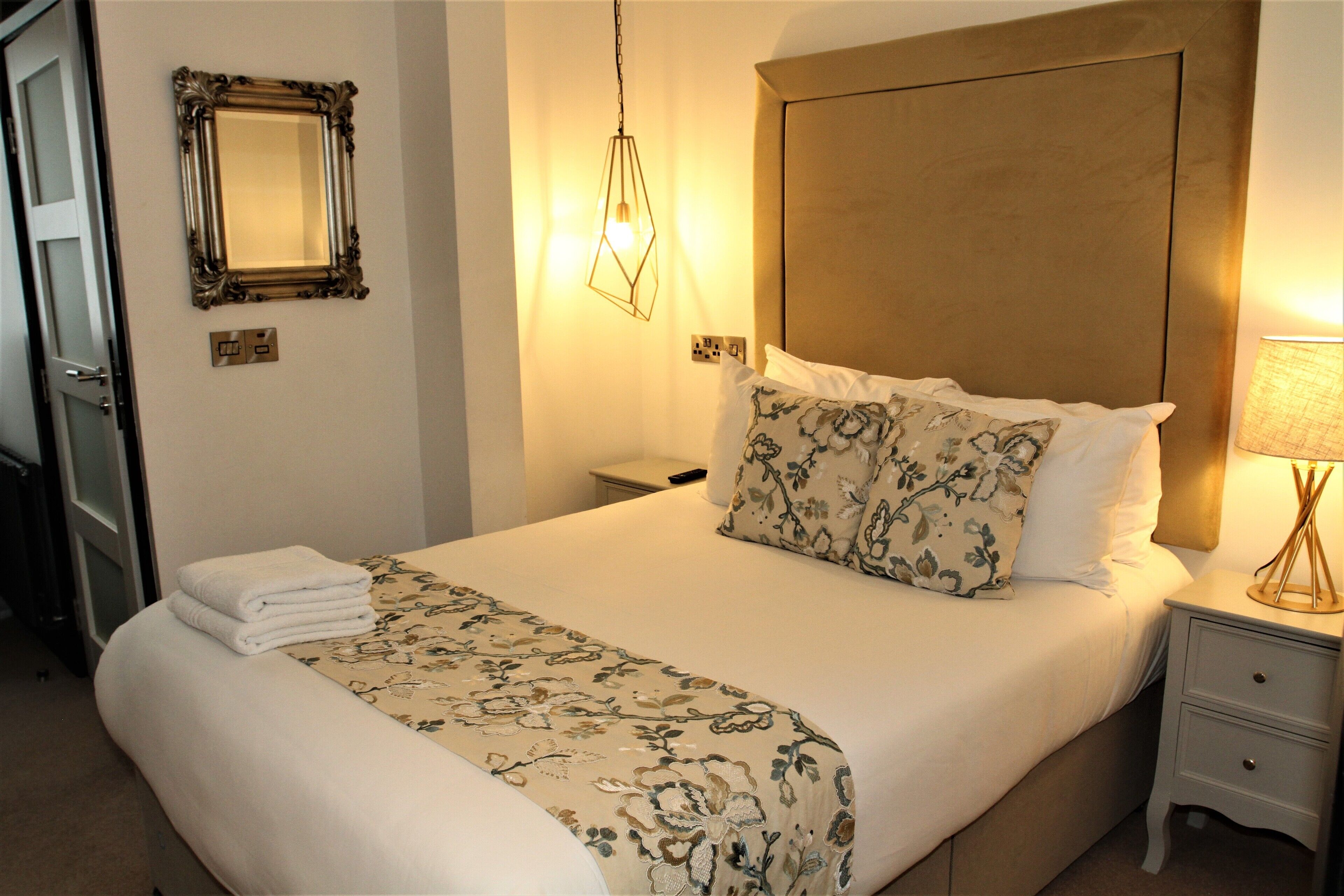 A neatly made bed with a floral bedspread, a bedside lamp, and a wall-mounted mirror.