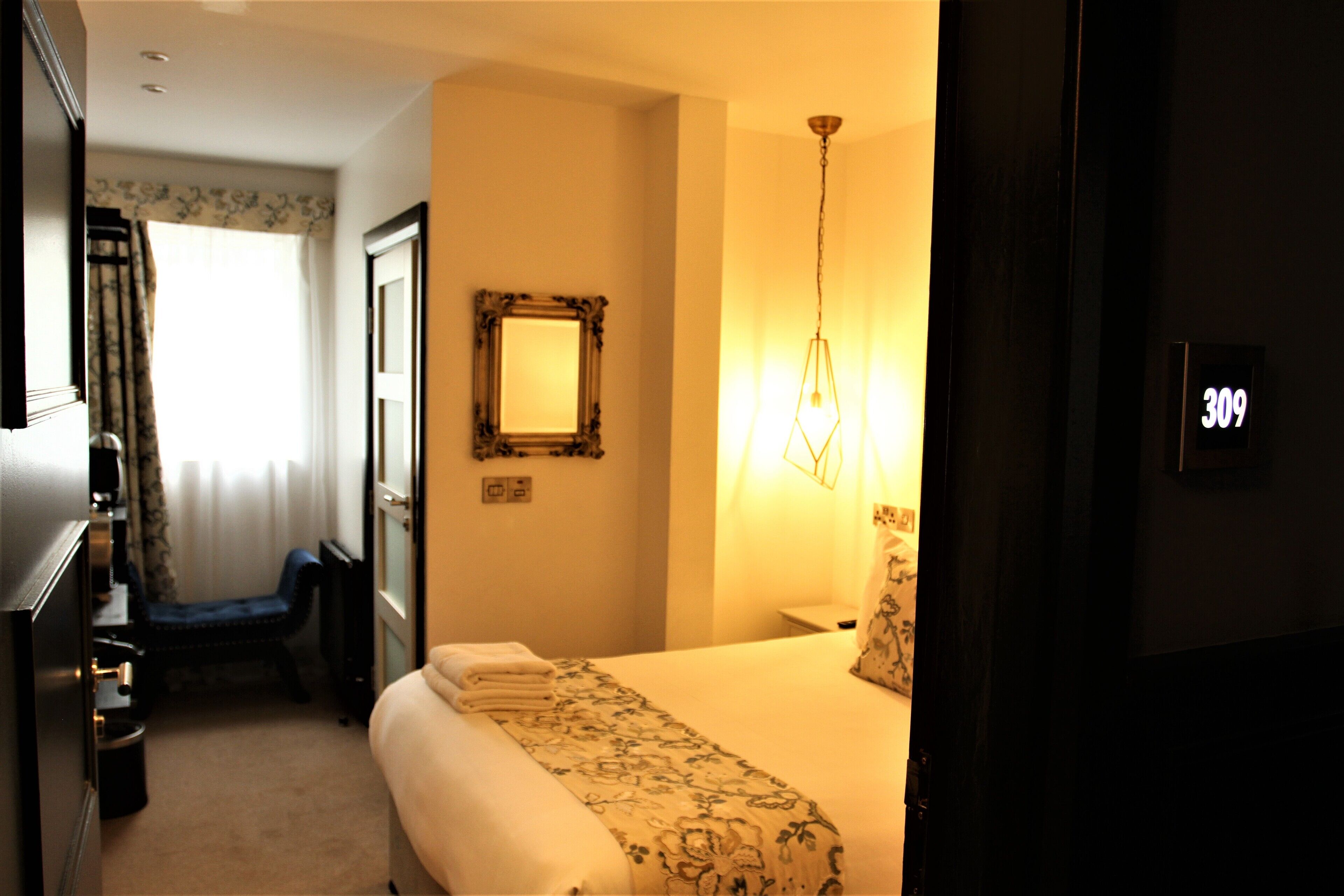A hotel room with a bed, a nightstand, a mirror, and a wall-mounted light fixture.