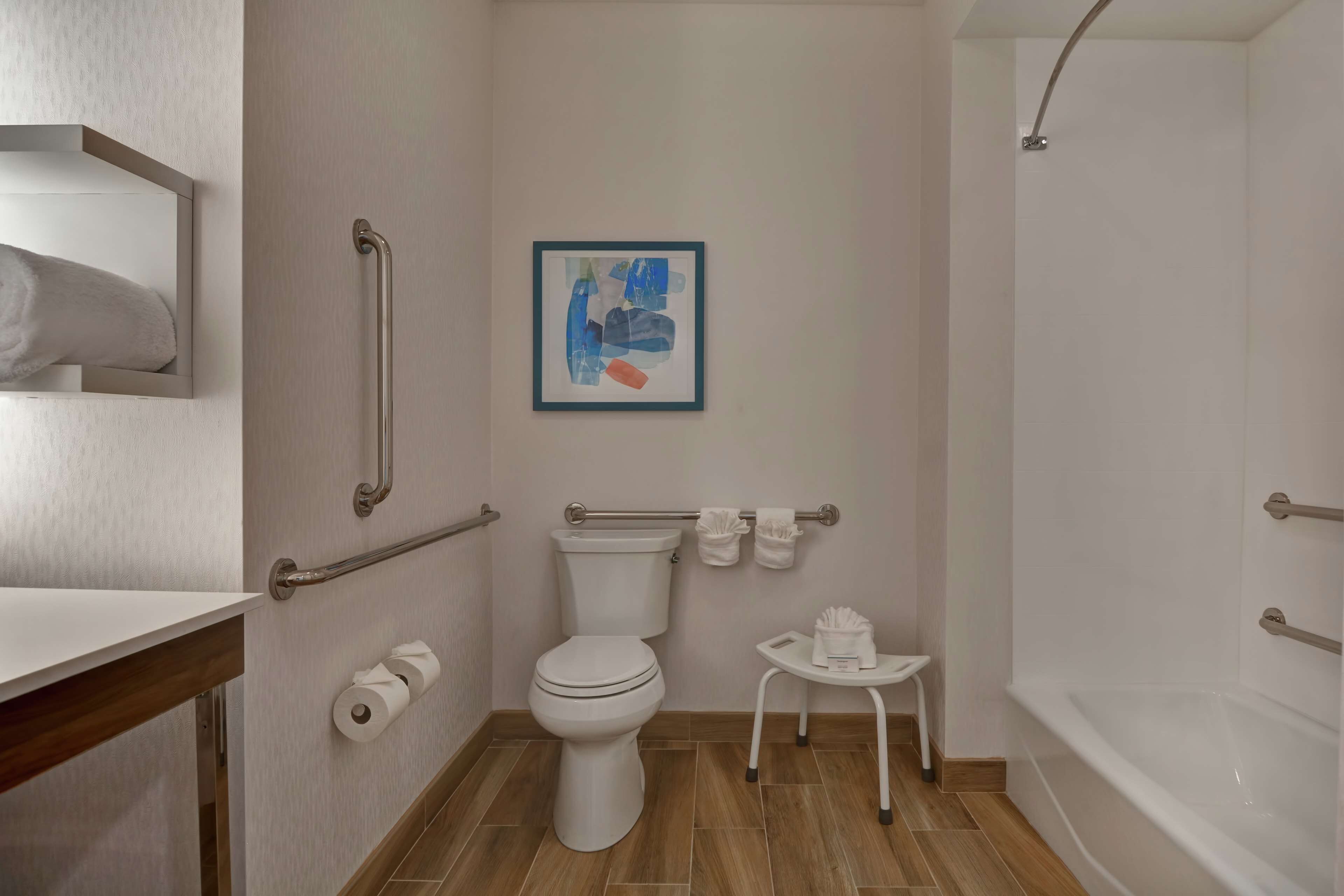 A bathroom with a toilet, handrails, a framed abstract painting, a small table with a tissue box, and a bathtub.