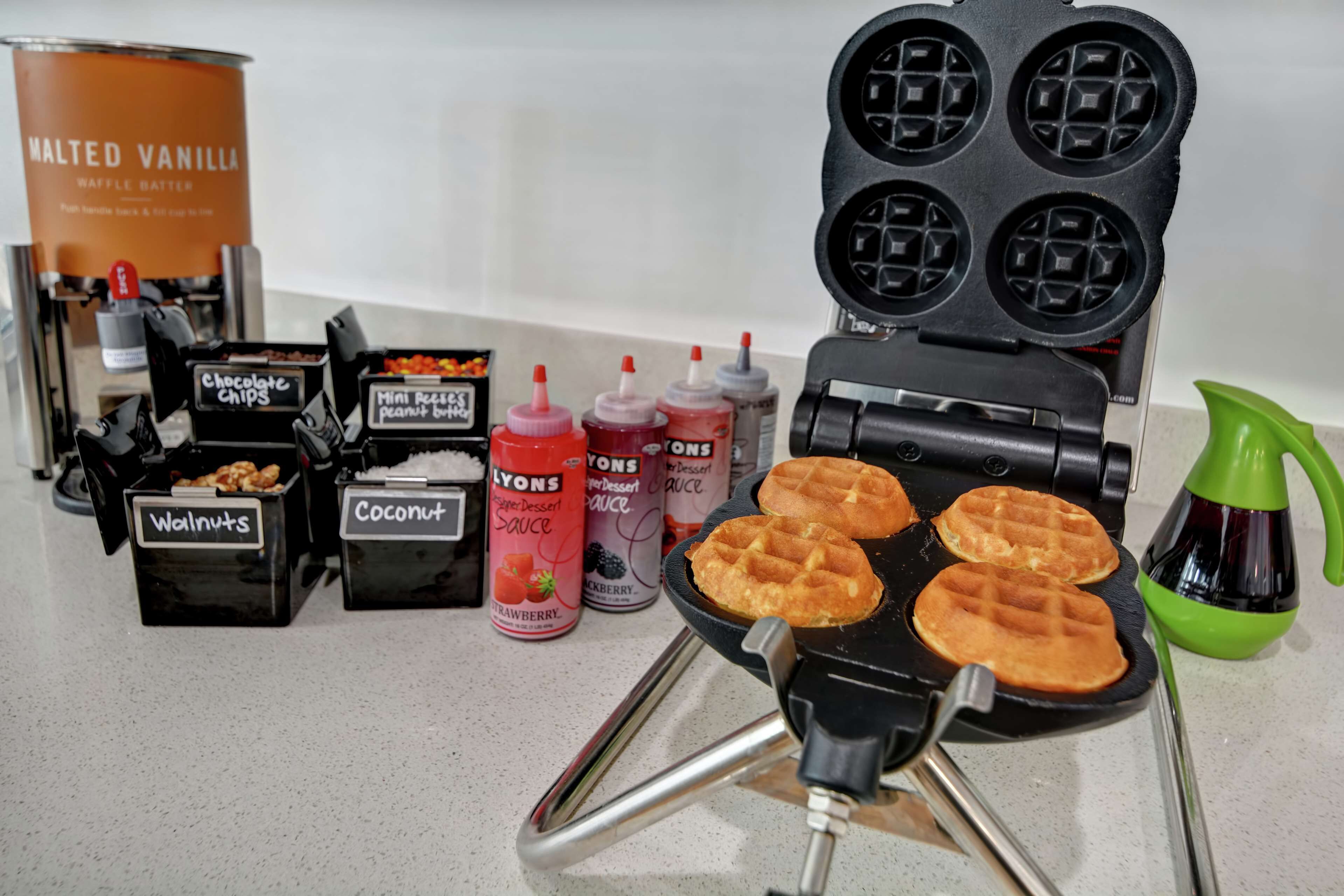 Ein Waffeleisen mit vier Waffeln, eine Dose Vanille-Waffel-Teig und verschiedene Toppings wie Schokoladenstückchen, Mini Reese’s Erdnussbutter, Walnüsse, Kokosraspeln und Sirups.