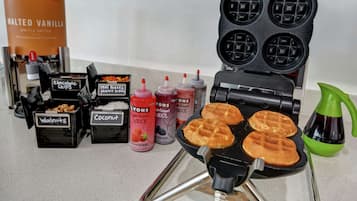 Ein Waffeleisen mit vier Waffeln, eine Dose Vanille-Waffel-Teig und verschiedene Toppings wie Schokoladenstückchen, Mini Reese’s Erdnussbutter, Walnüsse, Kokosraspeln und Sirups.