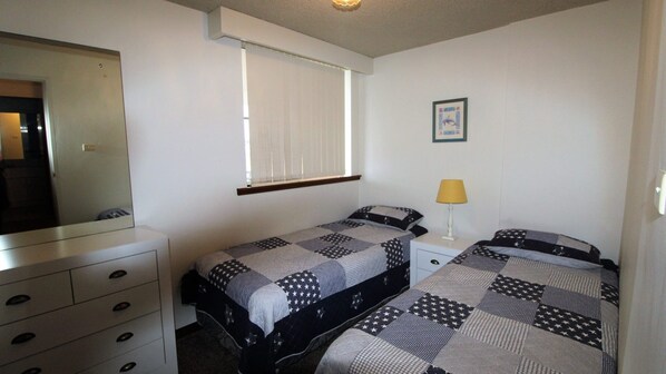 A bedroom with two beds, a dresser, a window with blinds, a lamp, and a framed picture on the wall.