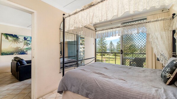 A bedroom with a bed, a chair, a window with curtains, and a view of trees and a building. - Ocean Court 8, 13 Lord Street (Port Macquarie)