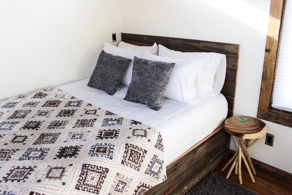 A neatly made bed with a patterned blanket and pillows, a small wooden stool, and a window with blinds. - Cliffside Cabin   # 2 (St Orderville)
