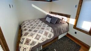 A bedroom with a sloped ceiling, skylights, a bed with patterned bedding, and a window with blinds. - Cliffside Cabin   # 2 (St Orderville)