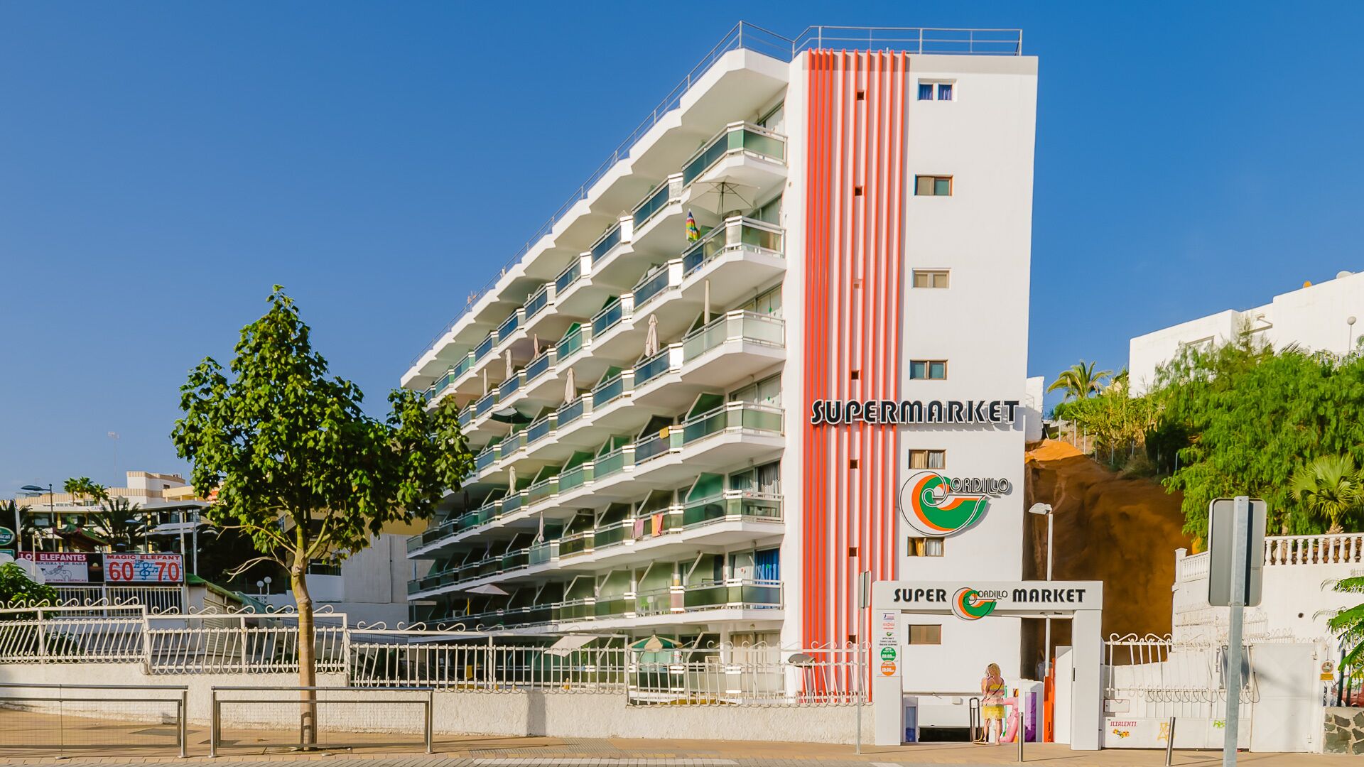 Ein mehrstöckiges Gebäude mit auffälliger, vertikaler, roter Streifen, Balkonen und einem Schild für 'Supermarket Cordillo'.