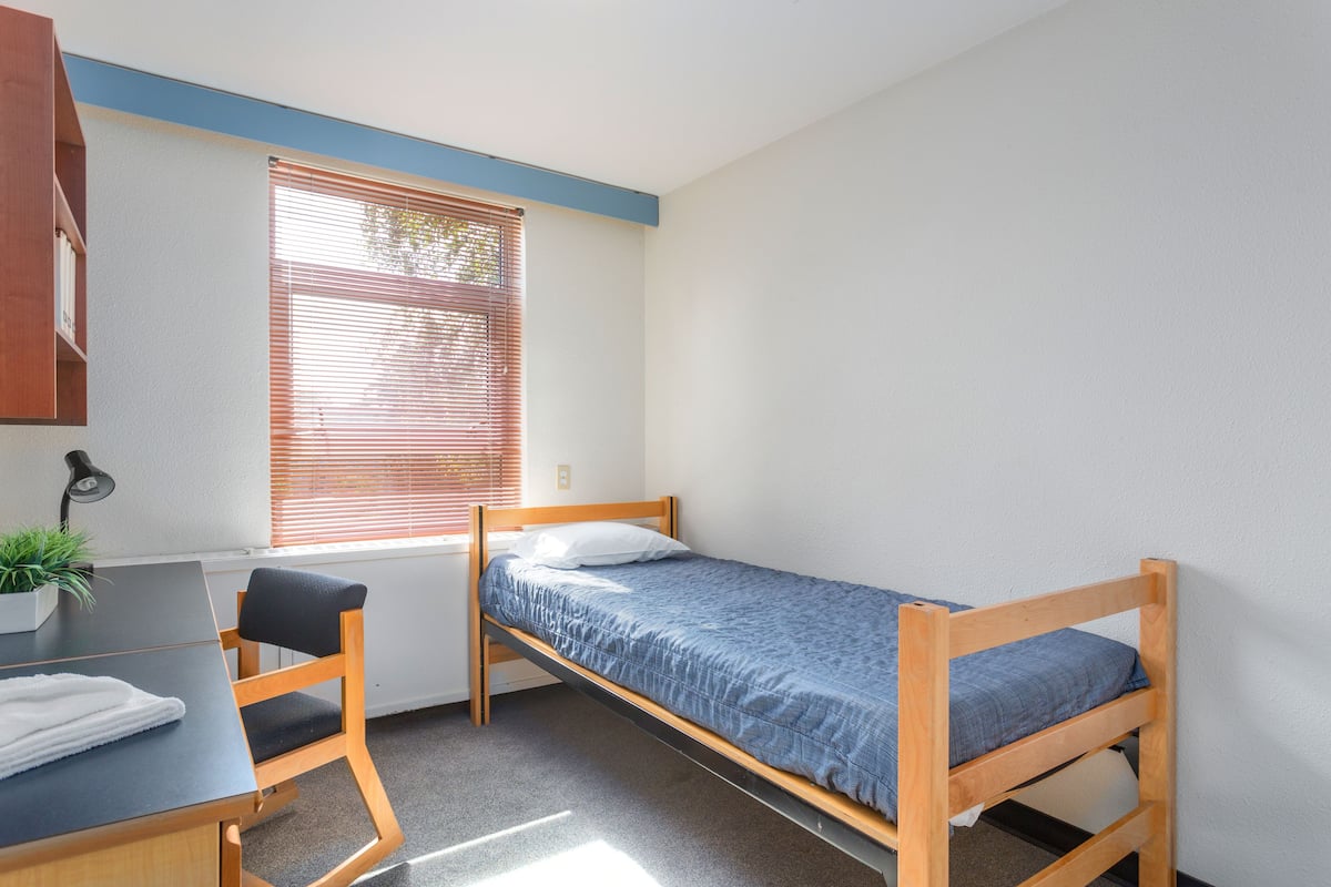 A dormitory room with a single bed, a desk, a chair, and a window with blinds.