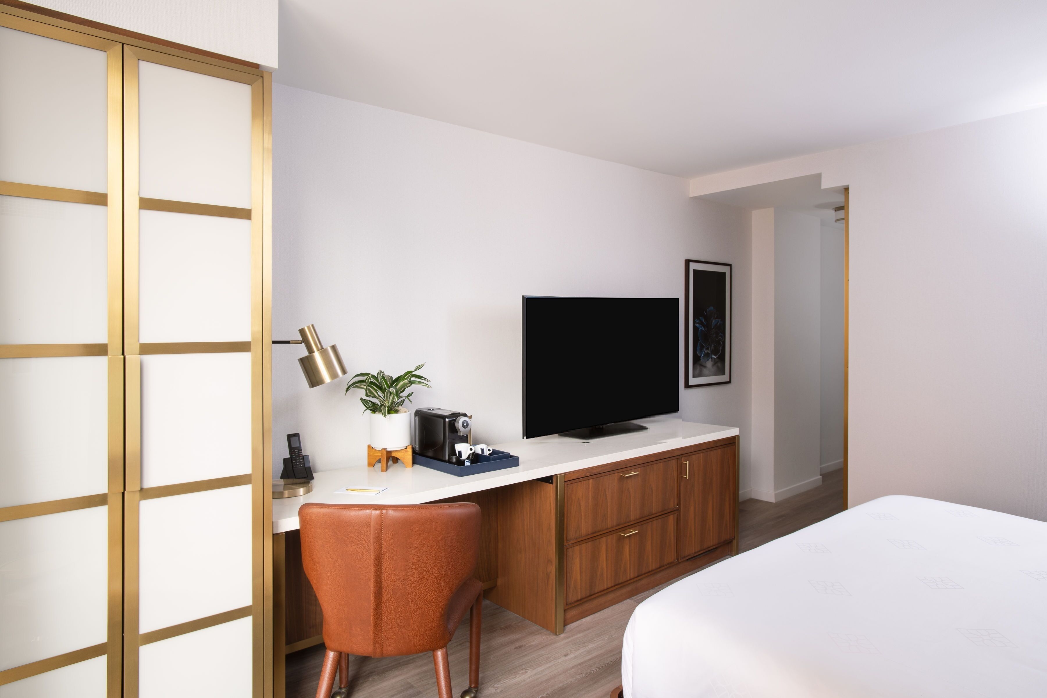 Moderni hotellihuone, jossa on sänky, työpöytä, jossa on televisio, tuoli ja pieni ruukkukasvi.